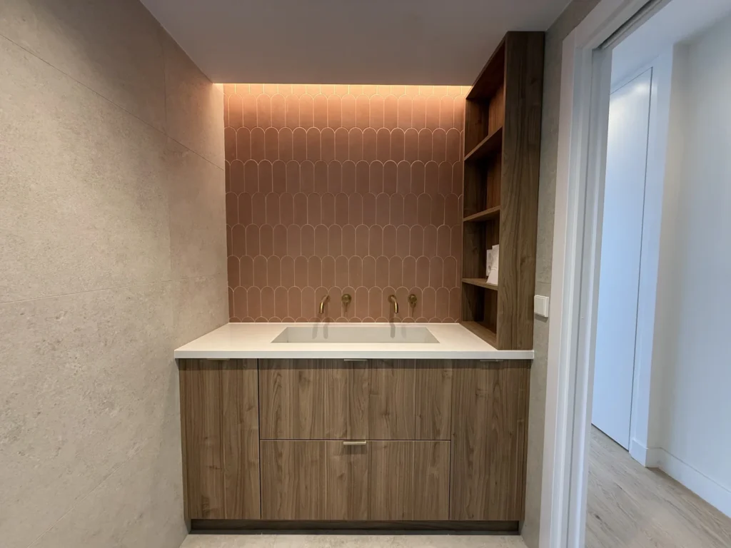 Vista lateral de reforma baño en Barrio de Tetuán: encimera con grifos dorados y estantería de madera junto a puerta.