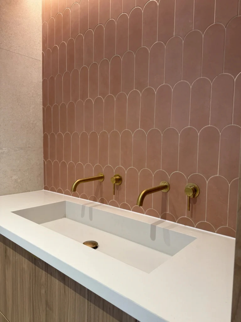 Interiorismo moderno en Barrio de Tetuán: baño con azulejos escamados en tonos rosados y grifos dorados, encimera blanca minimalista.